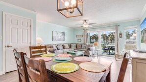 A dining area with a wooden table, chairs, and colorful placemats. There is a sofa and a window with a view of palm trees. - Bayfront Kontiki 3 Bedroom Condo by RedAwning (Rockport)