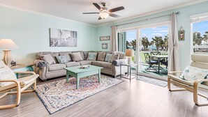 A spacious living room with a beige sofa, a coffee table, and a view of a balcony with palm trees. - Bayfront Kontiki 3 Bedroom Condo by RedAwning (Rockport)