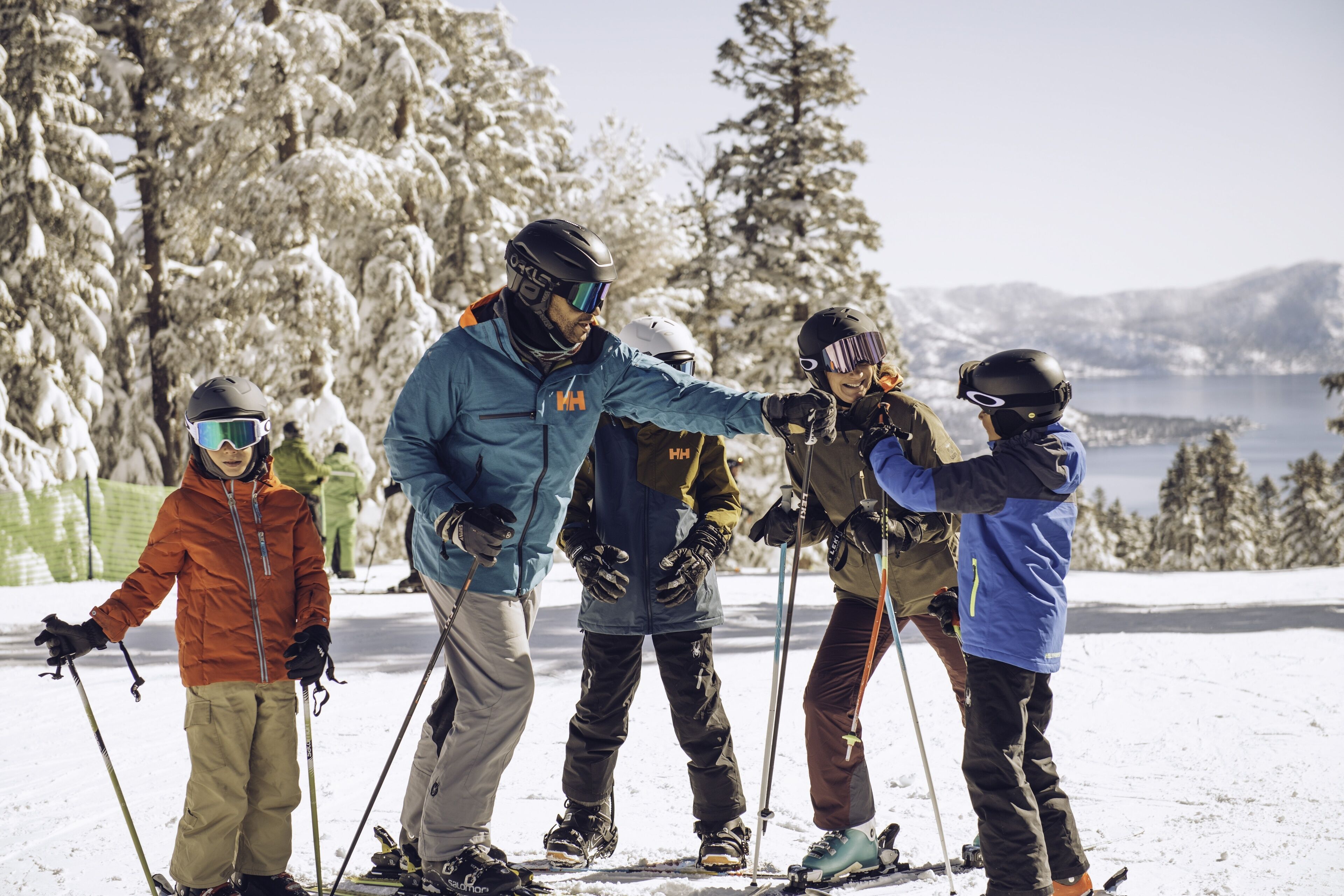 A group of skiers on a snowy slope with trees and a lake in the background.