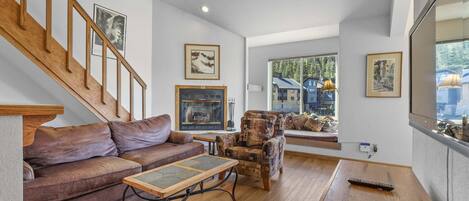 Un salón amplio con una escalera de madera, chimenea, sofá, sillón estampado y un ventanal con vistas al exterior.