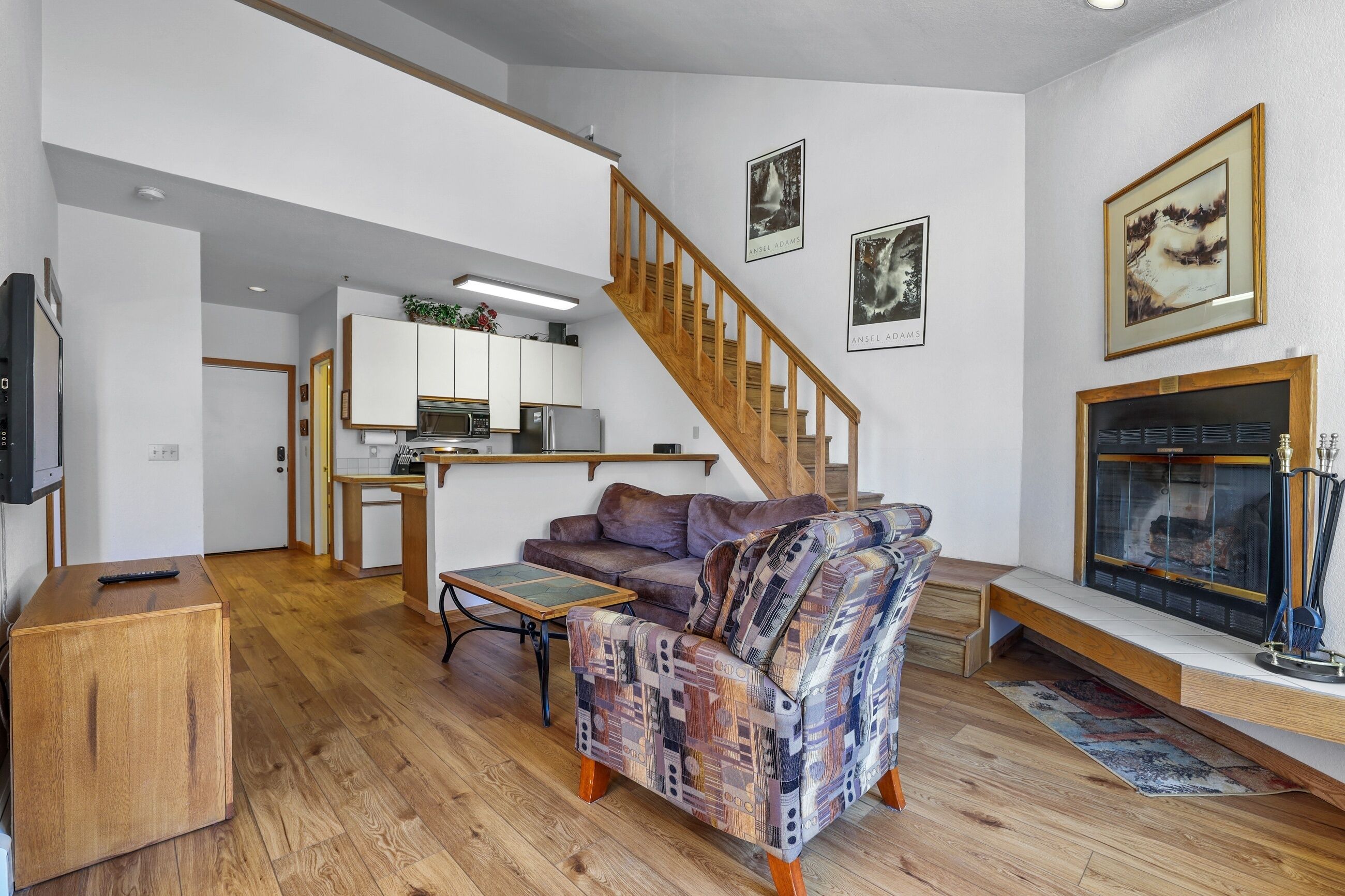 Una sala de estar con escalera de madera, chimenea y una cocina al fondo.