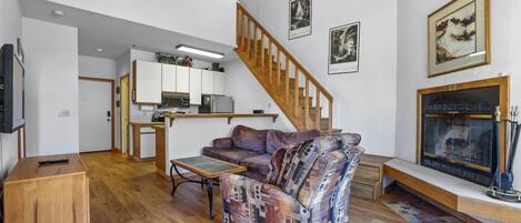 A living room with a wooden staircase, a fireplace, and a kitchen in the background.