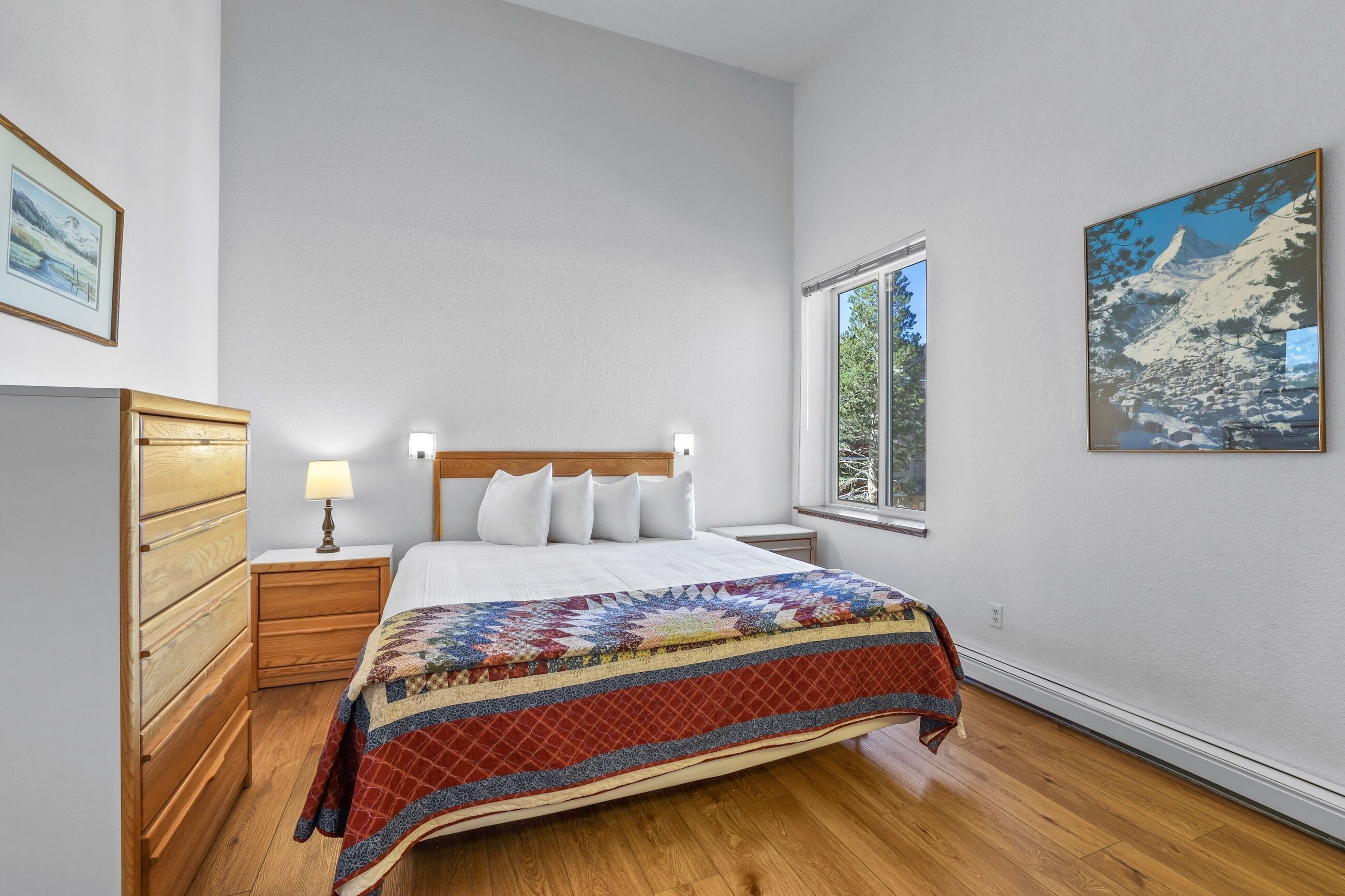 A bedroom with a wooden bed, a nightstand with a lamp, a window with a view of trees, and a painting on the wall.