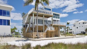 Een huis aan het strand met een houten terras, een balkon en een trap naar de bovenverdieping.