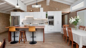 A modern kitchen with white cabinets, a central island with bar stools, and a dining area with wooden chairs and a table. - 4 Shelly Beach Road: 2  BR, 2  BA House in Port Macquarie , Sleeps 4 (Port Macquarie)