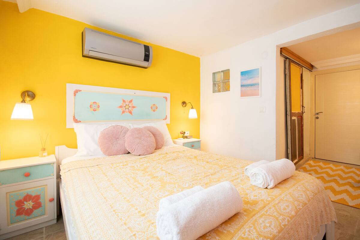 A hotel room with a yellow wall, a bed with a floral headboard, two bedside tables with lamps, and a door leading to a bathroom.
