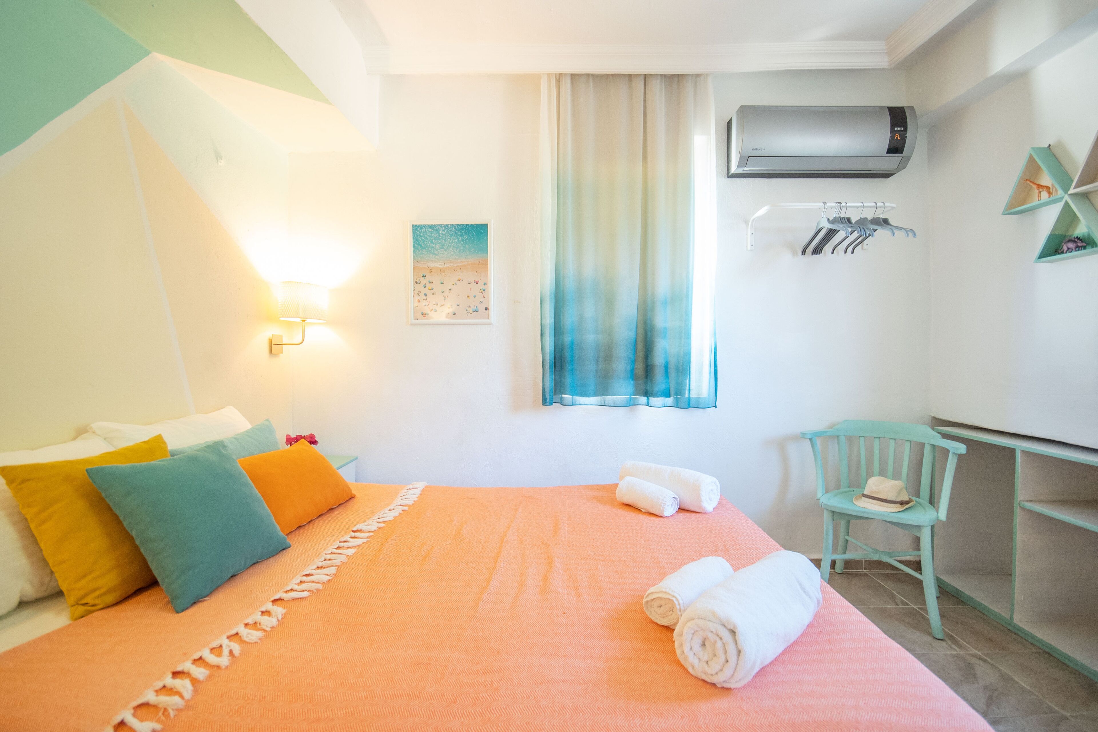 Un dormitorio con cama, almohadas, una ventana con cortinas, un equipo de aire acondicionado, una silla y un perchero en la pared con ropa.