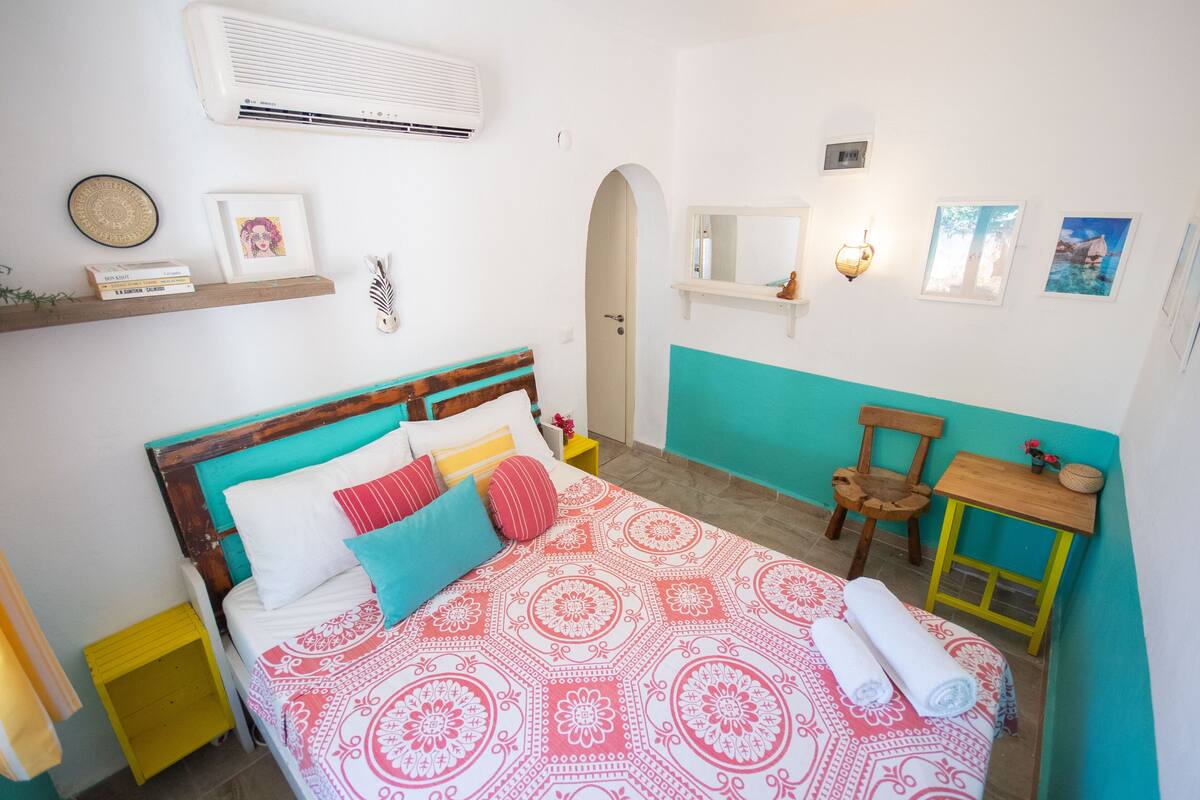 A bedroom with a bed, a chair, a small table, and a shelf with books and a picture frame.