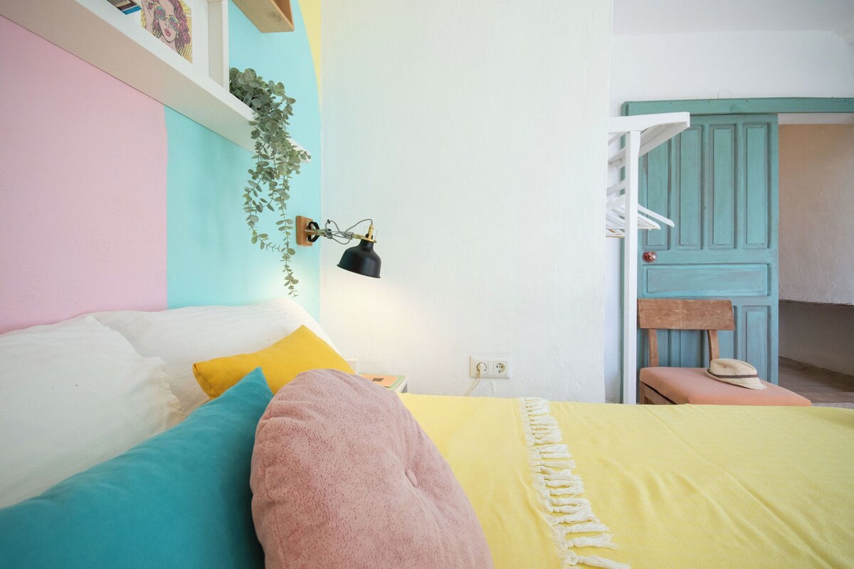 A bedroom with a bed, pillows, a bedside table, a lamp, a plant, and a wardrobe.