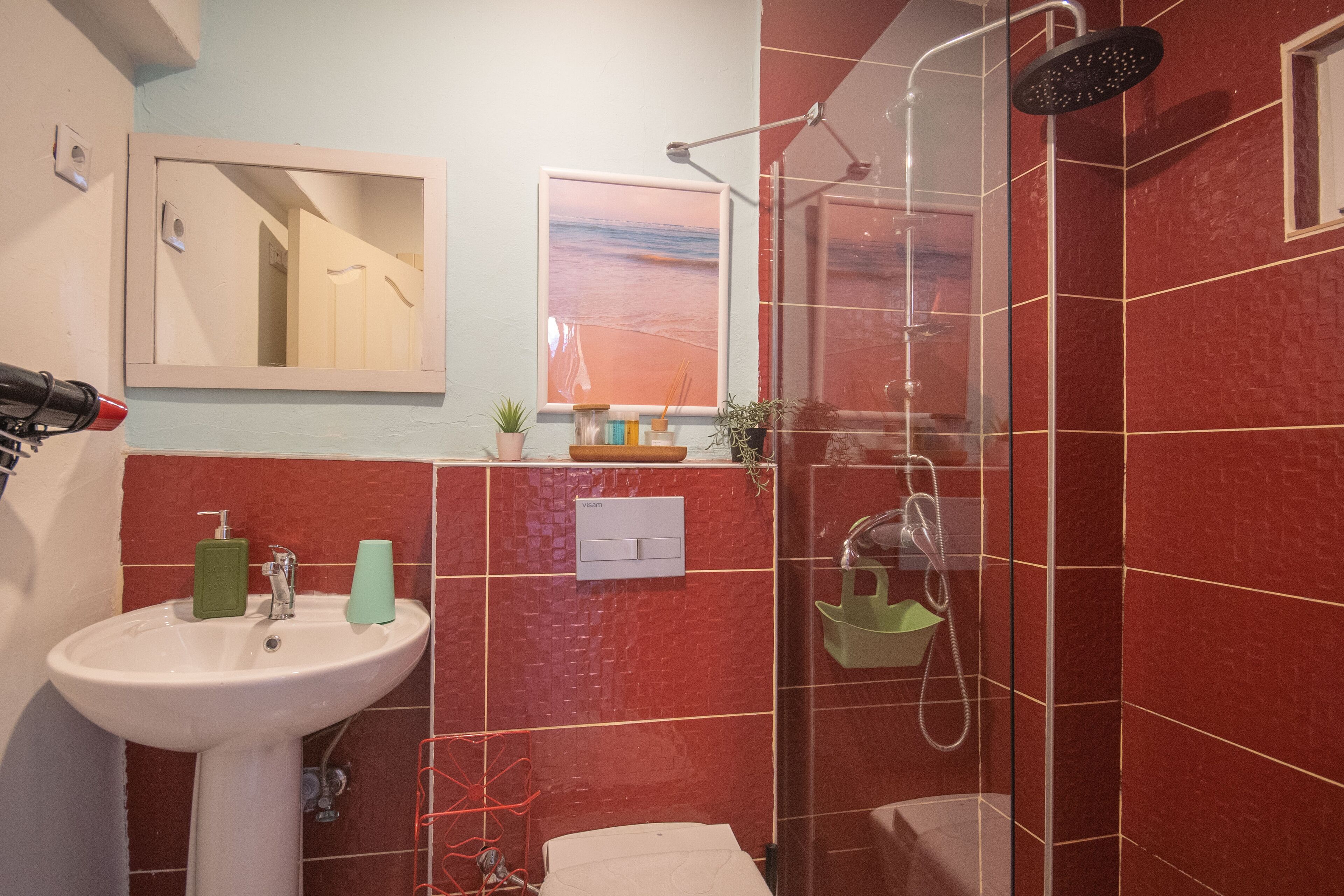 A bathroom with a white sink, red-tiled walls, a shower, and a framed picture of a beach.