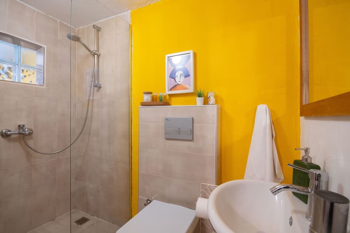 A bathroom with a shower, a sink, and a toilet. There is a yellow wall and a framed picture above the sink.