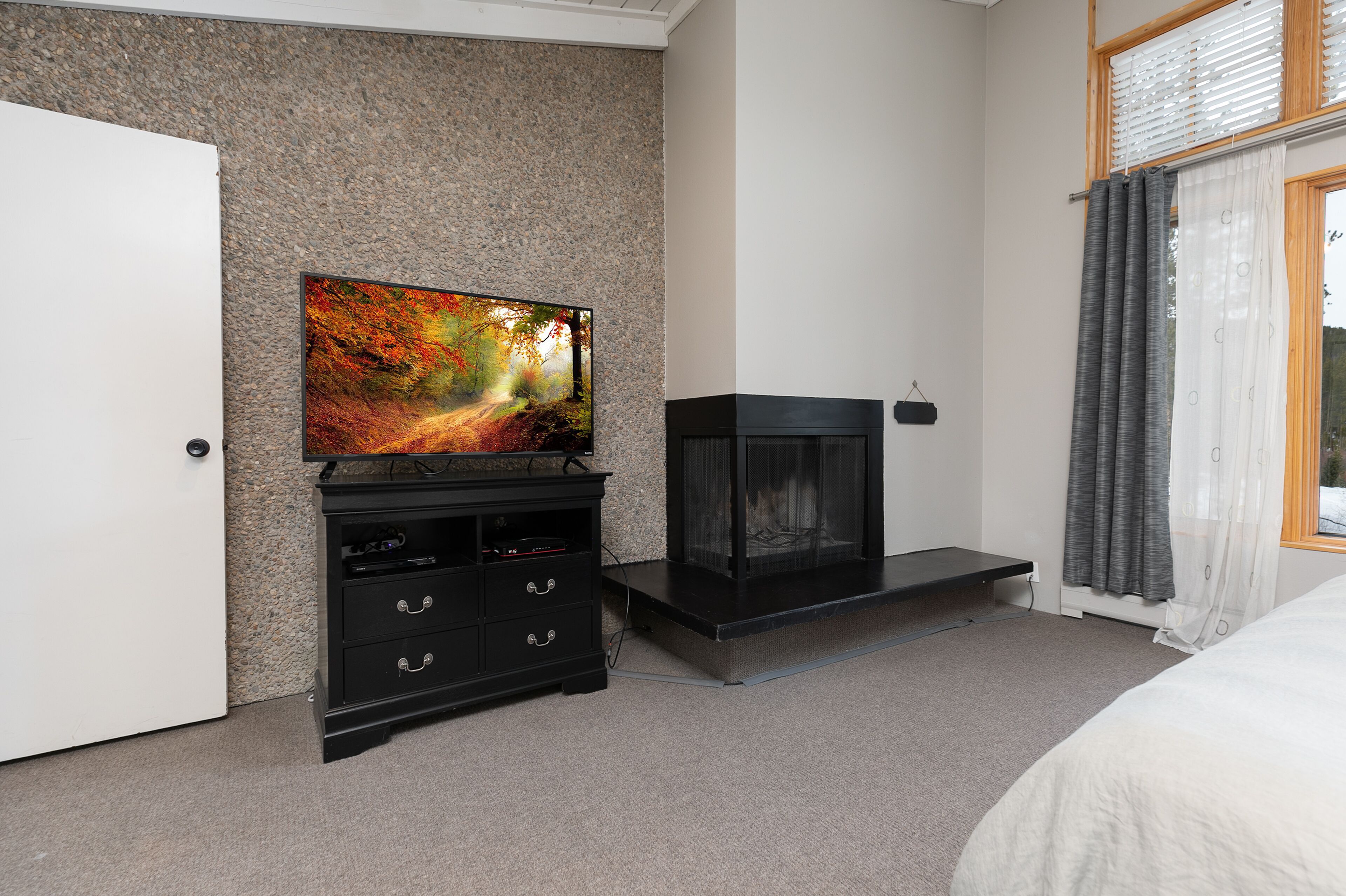 Un dormitorio con una televisión sobre un soporte negro, una chimenea, una puerta y una ventana con cortinas.