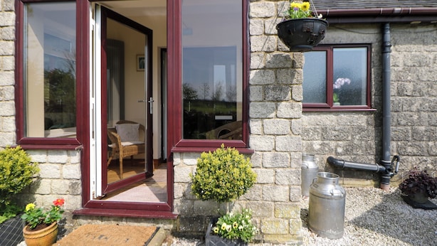 Una casa de piedra con un marco de puerta y ventanas rojo, un pequeño jardín con plantas en macetas y un depósito de agua metálico.