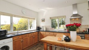 Una cocina moderna con armarios de madera, lavadora integrada, zona de comedor con mesa y sillas, y ventanas amplias con vistas a zonas verdes.