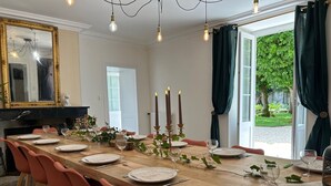Een eetkamer met een lange houten tafel, gedekt voor zes personen, omringd door rode stoelen. Er staan kaarsen op tafel en een kroonluchter hangt aan het plafond.