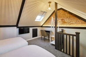 A bedroom with two beds, a desk, and a chair. There is a skylight and a stone wall.