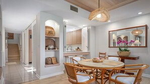 A dining area with a round table and wicker chairs, a kitchen with white cabinets, and a staircase leading to another level. - The Islands M2 Oasis at Mauna Lani Resort (Kamuela)