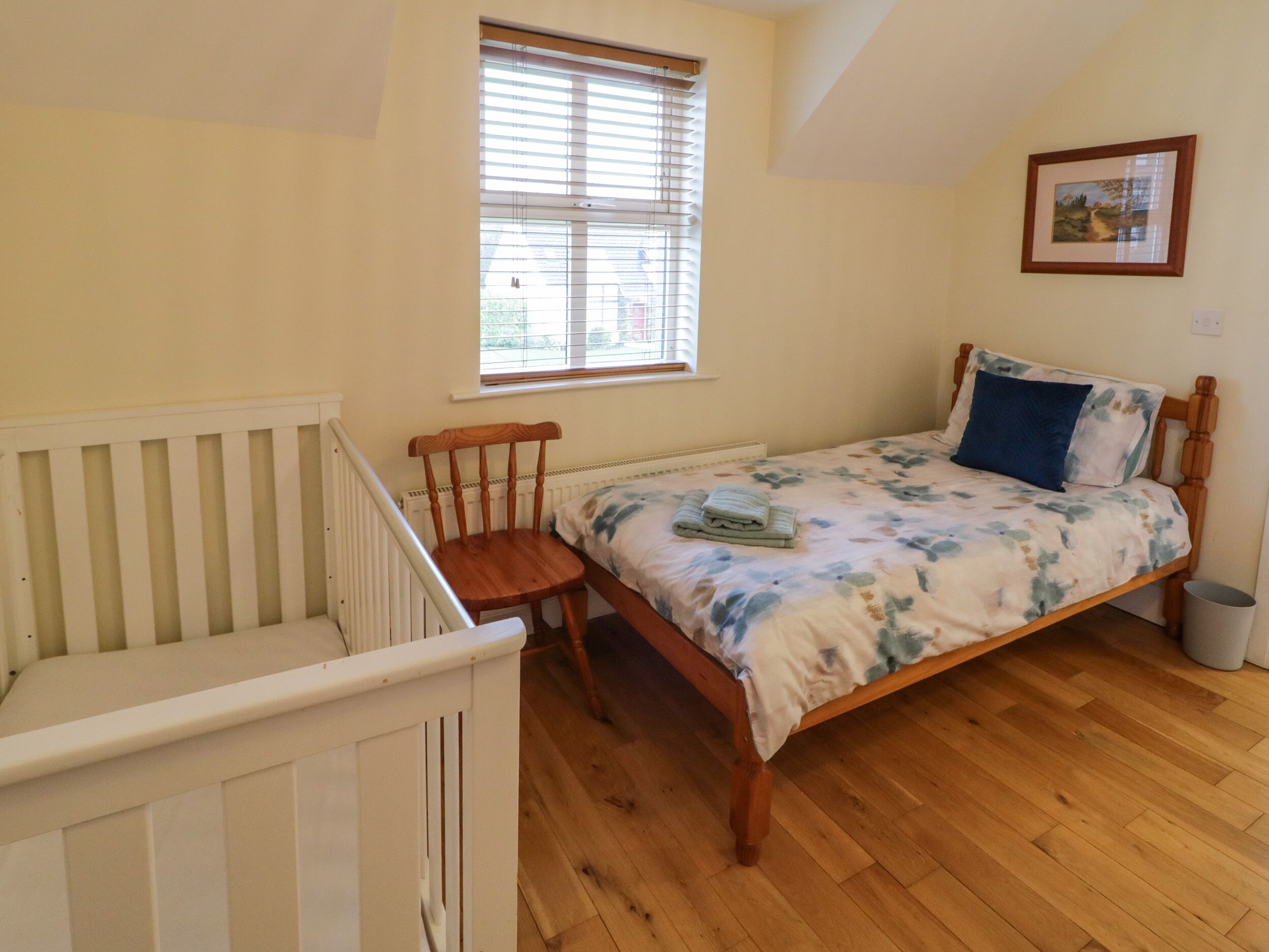 A bedroom with a wooden bed, a crib, a window with blinds, and a framed picture on the wall.