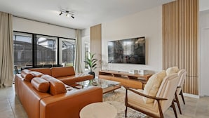 A modern living room with a leather sofa, a glass coffee table, a wooden TV stand, and a large flat-screen TV mounted on the wall.