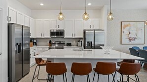 A modern kitchen with white cabinetry, stainless steel appliances, a central island with bar stools, pendant lighting, and a framed artwork on the wall.