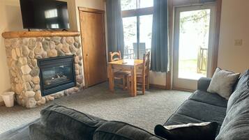Una sala de estar con chimenea de piedra, un televisor montado encima y un sofá gris oscuro, junto con una zona de comedor con mesa y sillas.