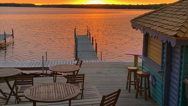 A wooden deck with tables and chairs overlooking a calm body of water at sunset. - Manhattan Beach Lodge (Crosslake)