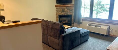 A living room with a stone fireplace, a flat-screen TV, and a large window with curtains.