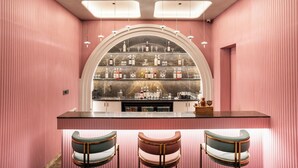 Zona de bar con una estantería curva que exhibe botellas y copas, una encimera con utensilios de bar y tres taburetes.