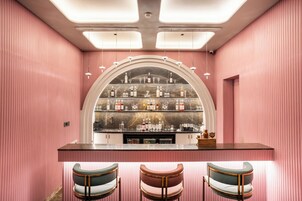 Uma área de bar com uma estante curva exibindo garrafas e copos, um balcão com utensílios de bar e três banquetas.