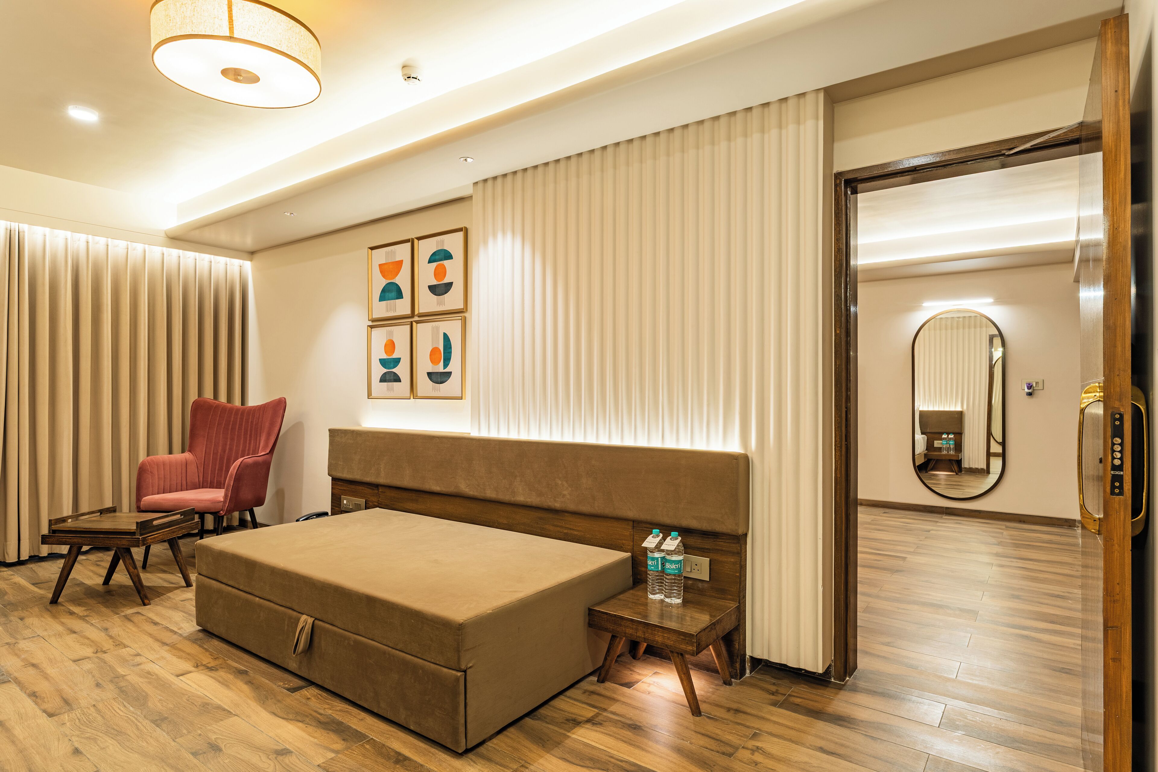 Een hotelkamer met een groot bed, een houten nachtkastje, een rode stoel, een kleine tafel, een waterfles en een spiegel.