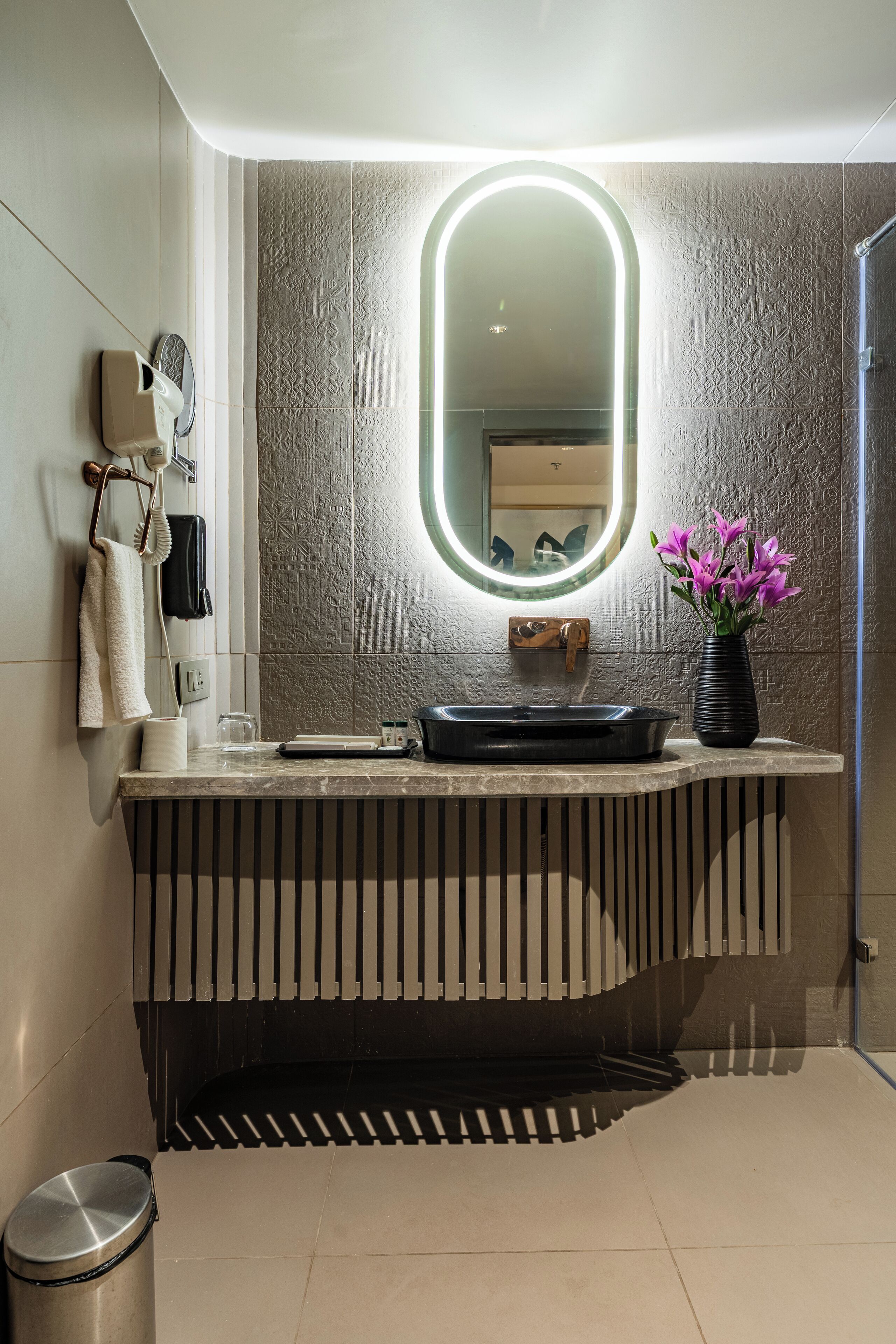 A modern bathroom with a vanity, a mirror with a light, a trash bin, and a vase with flowers.