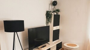 Ein modernes Wohnzimmer mit einem Flachbildfernseher auf einer weißen Konsole, einer Stehlampe, einem runden Beistelltisch und einer Pflanze an der Wand.