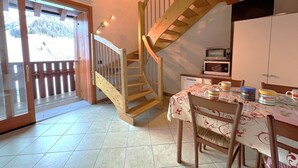 A kitchen with a wooden staircase, a dining table with chairs, and a view of a snowy landscape through a glass door. - San Simone rentals - VALS30 (Valleve)