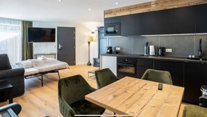 A modern hotel room with a bed, a TV, a desk, a chair, a table, a kitchen area with cabinets and appliances, and a window with curtains. - Love the Alps Apartments (Reith bei Seefeld)