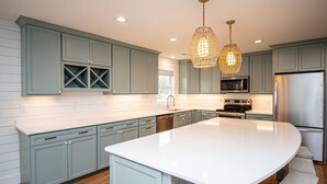 Una cocina moderna con armarios azul claro, una isla blanca, electrodomésticos de acero inoxidable y lámparas colgantes de mimbre.