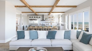 Ein modernes Wohnzimmer mit einer großen weißen Couch, Holzbalken, einer Küche im Hintergrund und Blick aufs Meer.