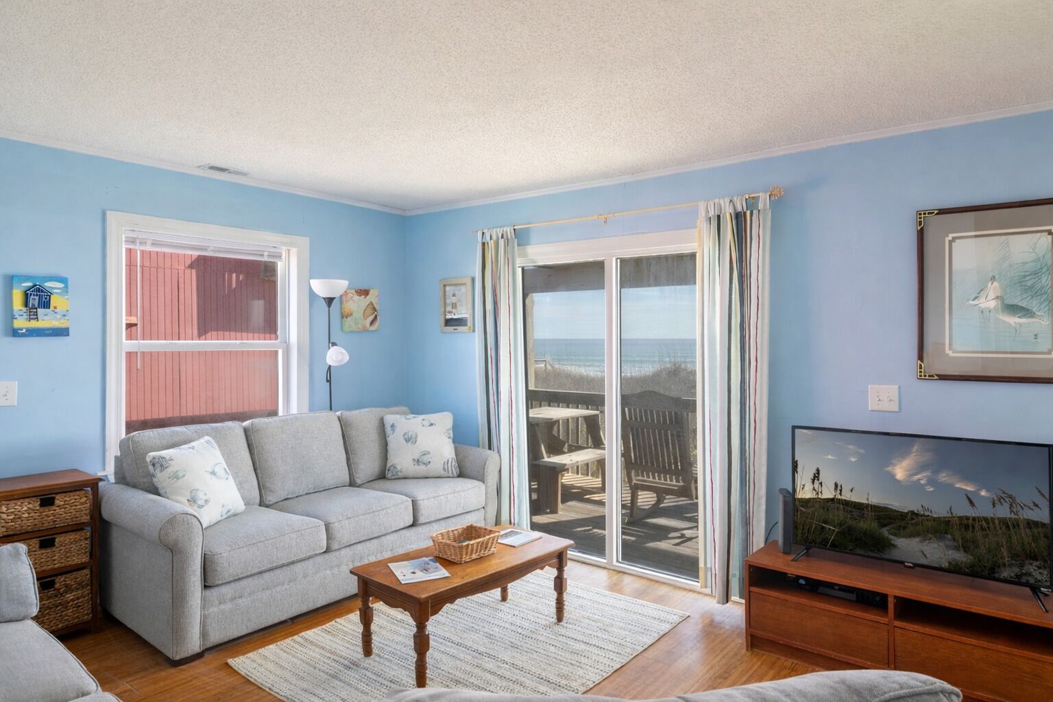 Una sala de estar con sofá gris, mesa de centro de madera, mueble para televisor y vista a la playa a través de grandes puertas correderas de vidrio.