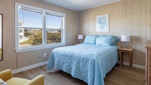 A bedroom with a large window, wooden panel walls, a bed with blue bedding, two bedside tables with lamps, and a framed picture on the wall. - Sassy Seagull West, Oceanfront Duplex in Emerald Isle, NC! (Emerald Isle)