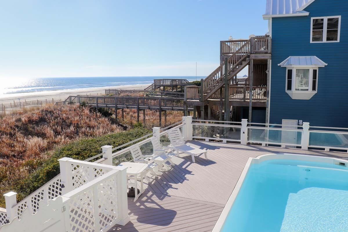 Zona de piscina con vista a la playa, terraza de madera y un edificio azul con balcón.
