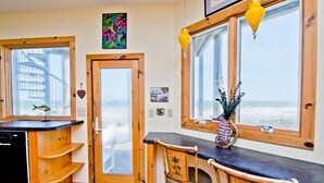 A cozy room with wooden furniture, a desk, and a chair. There are two windows with curtains, a door, and a small shelf with a plant.