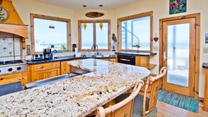 A kitchen with granite countertops, wooden cabinets, and a large island. There are two windows with views of the outside, and a door leading to a balcony.