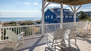 Une terrasse en bois avec des chaises Adirondack blanches donnant sur une plage et une maison bleue.