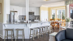 Une cuisine spacieuse avec des meubles blancs, un îlot central en granit doté de tabourets, et un coin repas avec des chaises et une table en bois.