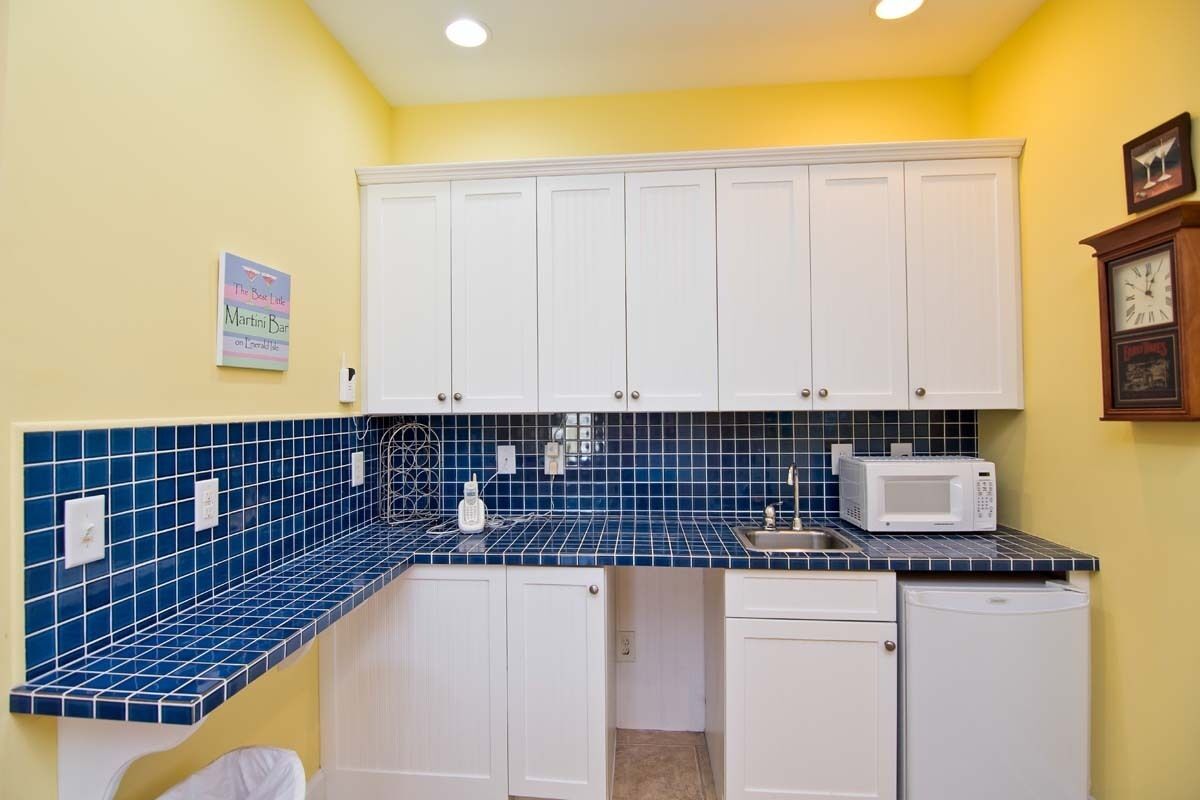 A compact kitchen with white cabinets, a blue tiled backsplash, a microwave, and a clock on the wall.