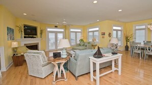 A living room with a fireplace, a television, a sofa, a coffee table, and a dining area with chairs and a table. - Sea Notes, Almost Oceanfront House in Emerald Isle, NC! (Emerald Isle)
