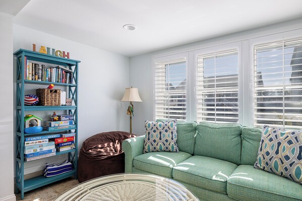 A cozy living room with a teal bookshelf, a green sofa, and a wicker chair. - Sea Star, Oceanside House in Atlantic Beach, NC! (Atlantic Beach)
