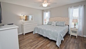 Een slaapkamer met een bed, een dressoir, een spiegel, een plafondventilator en een raam met gordijnen.