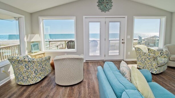Een ruime woonkamer met een groot raam dat uitkijkt op het strand, een hoge plafond en een decoratief element in het plafond.