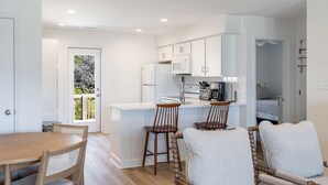 A modern kitchen with white cabinets, a central island, and a dining area with wooden chairs and a table. - Seabatical East, Almost Oceanfront Duplex in Emerald Isle, NC! (Emerald Isle)
