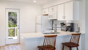 A modern kitchen with white cabinetry, a central island with bar stools, and a view of the outdoors through a door. - Seabatical East, Almost Oceanfront Duplex in Emerald Isle, NC! (Emerald Isle)
